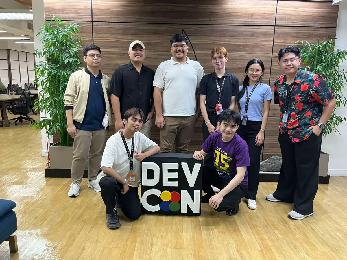 Jumpstart interns and mentors together, kneeling and standing around a handmade DEVCON sign