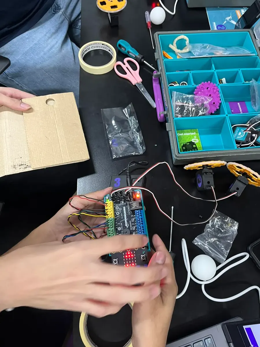 Hands wiring a Hummingbird robotics board with servos, tape, and cardboard on a workbench